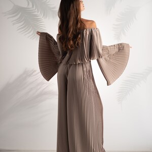 May include: A woman wearing a light brown off-the-shoulder top and matching wide-leg pants. The top and pants are made of pleated fabric.