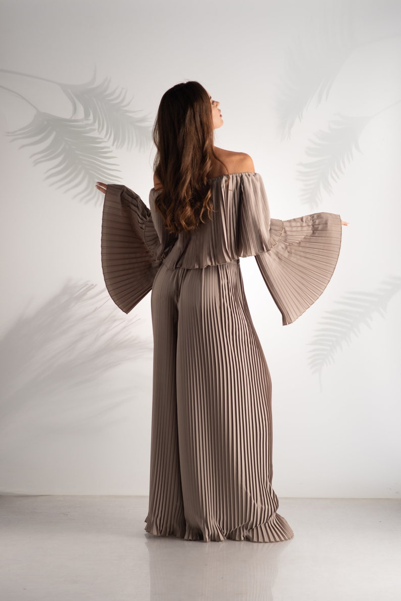 May include: A woman wearing a light brown off-the-shoulder top and matching wide-leg pants. The top and pants are made of pleated fabric.