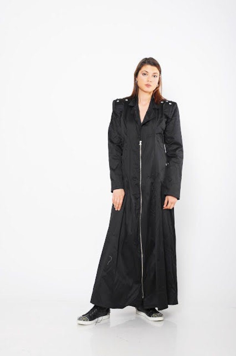 May include: A long black coat with a zipper closure and a stand-up collar. The coat has a slightly flared silhouette and features decorative accents on the shoulders.