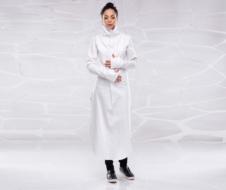May include: A white long-sleeved tunic with a high collar and long sleeves. The tunic has a relaxed fit and falls to the ankles. The model is wearing black pants and black shoes.