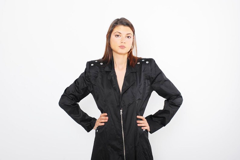 May include: A woman wearing a black jacket with silver accents and a zipper closure. She is standing with her arms crossed and looking directly at the camera.