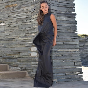 May include: A sleeveless black dress with a high neckline and a flowing skirt. The dress appears to be made of a smooth, possibly satin-like fabric. The model is standing next to a stone wall.