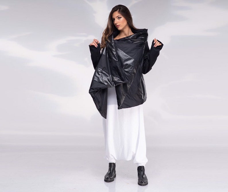 K&ouml;nnte beinhalten: Eine schwarze gesteppte Jacke mit einem gro&szlig;en, asymmetrischen Kragen. Die Jacke hat eine gl&auml;nzende, fast metallische Oberfl&auml;che. Das Model tr&auml;gt eine wei&szlig;e Hose und schwarze Stiefel.