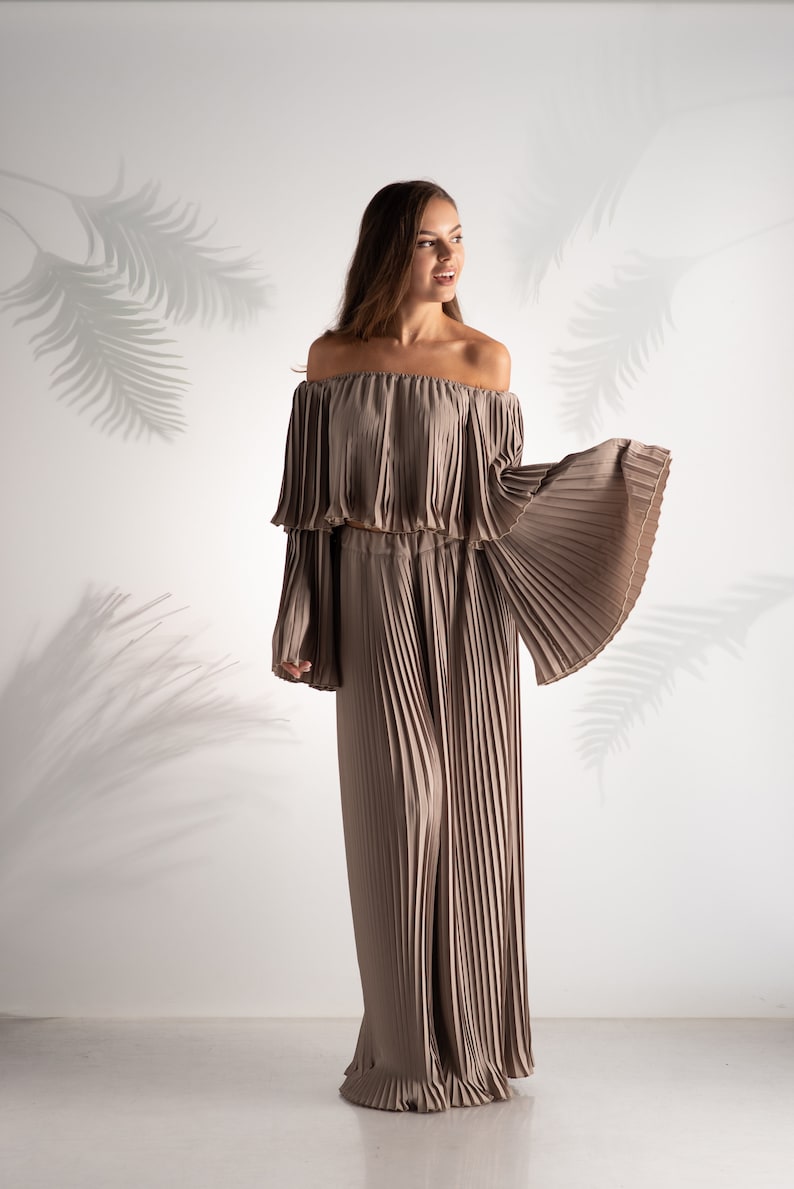 May include: A woman wearing a light brown off-the-shoulder top and matching wide-leg pants. Both pieces are made of pleated fabric.
