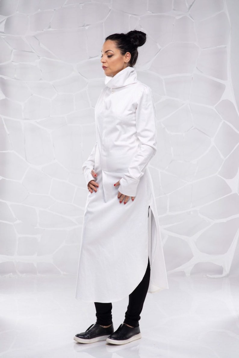 May include: A white long-sleeved shirt with a high collar and a long, flowing hem. The shirt has a side slit and is worn with black leggings and black shoes.