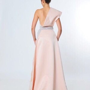 May include: A one-shoulder, blush pink maxi dress with a silver sash. The dress has a flowing skirt and a fitted bodice.
