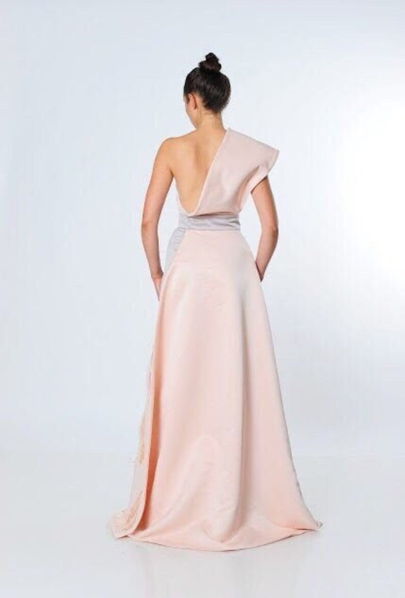 May include: A one-shoulder, blush pink maxi dress with a silver sash. The dress has a flowing skirt and a fitted bodice.