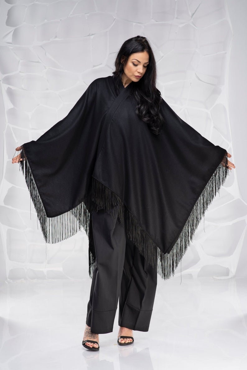 Puede incluir: Un poncho negro con flecos. El poncho se lleva sobre pantalones negros y la modelo lleva tacones negros.