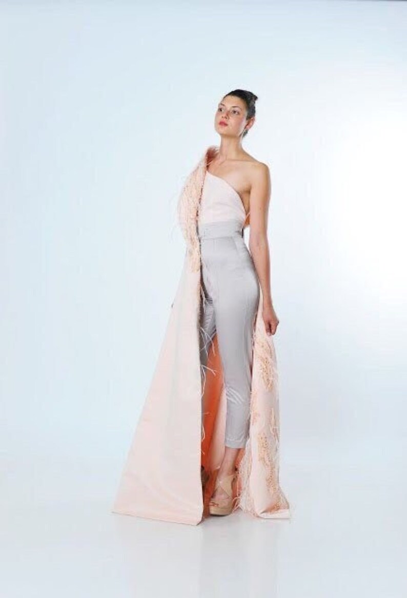 May include: A woman wearing a gray jumpsuit with a pink and peach feathered cape. The cape is draped over one shoulder and has a long train.