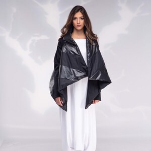 K&ouml;nnte beinhalten: Eine schwarze gesteppte Jacke mit einem langen, asymmetrischen Saum. Die Jacke hat einen hohen Kragen und einen Rei&szlig;verschluss. Das Model tr&auml;gt die Jacke &uuml;ber einem wei&szlig;en Oberteil und einer wei&szlig;en Hose.