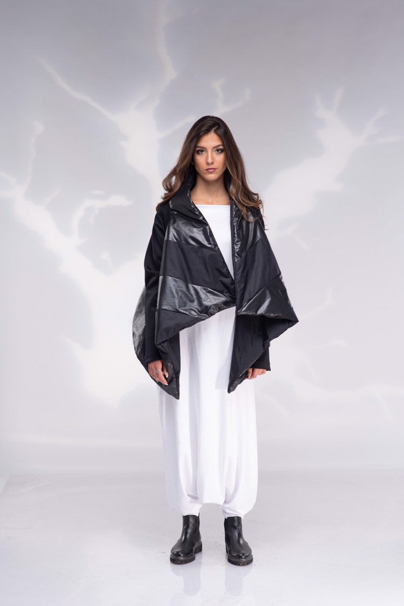 K&ouml;nnte beinhalten: Eine schwarze gesteppte Jacke mit einem langen, asymmetrischen Saum. Die Jacke hat einen hohen Kragen und einen Rei&szlig;verschluss. Das Model tr&auml;gt die Jacke &uuml;ber einem wei&szlig;en Oberteil und einer wei&szlig;en Hose.