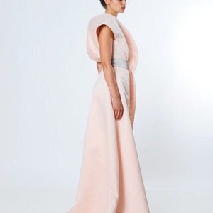 May include: A woman wearing a light pink dress with a large, draped, asymmetrical skirt and a fitted bodice. The dress has a silver belt at the waist.