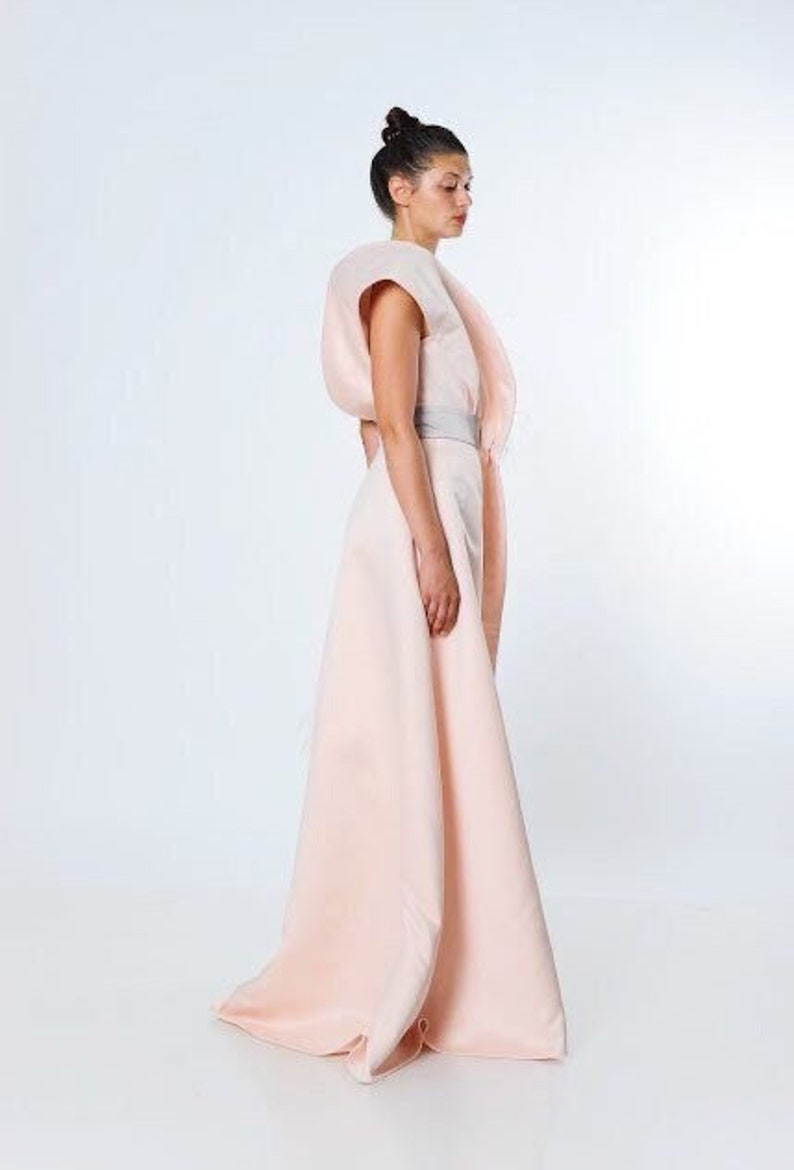 May include: A woman wearing a light pink dress with a large, draped, asymmetrical skirt and a fitted bodice. The dress has a silver belt at the waist.