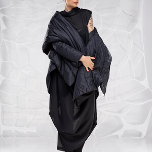 May include: A woman wearing a black, quilted jacket with a long, flowing skirt. She is standing in front of a white background.