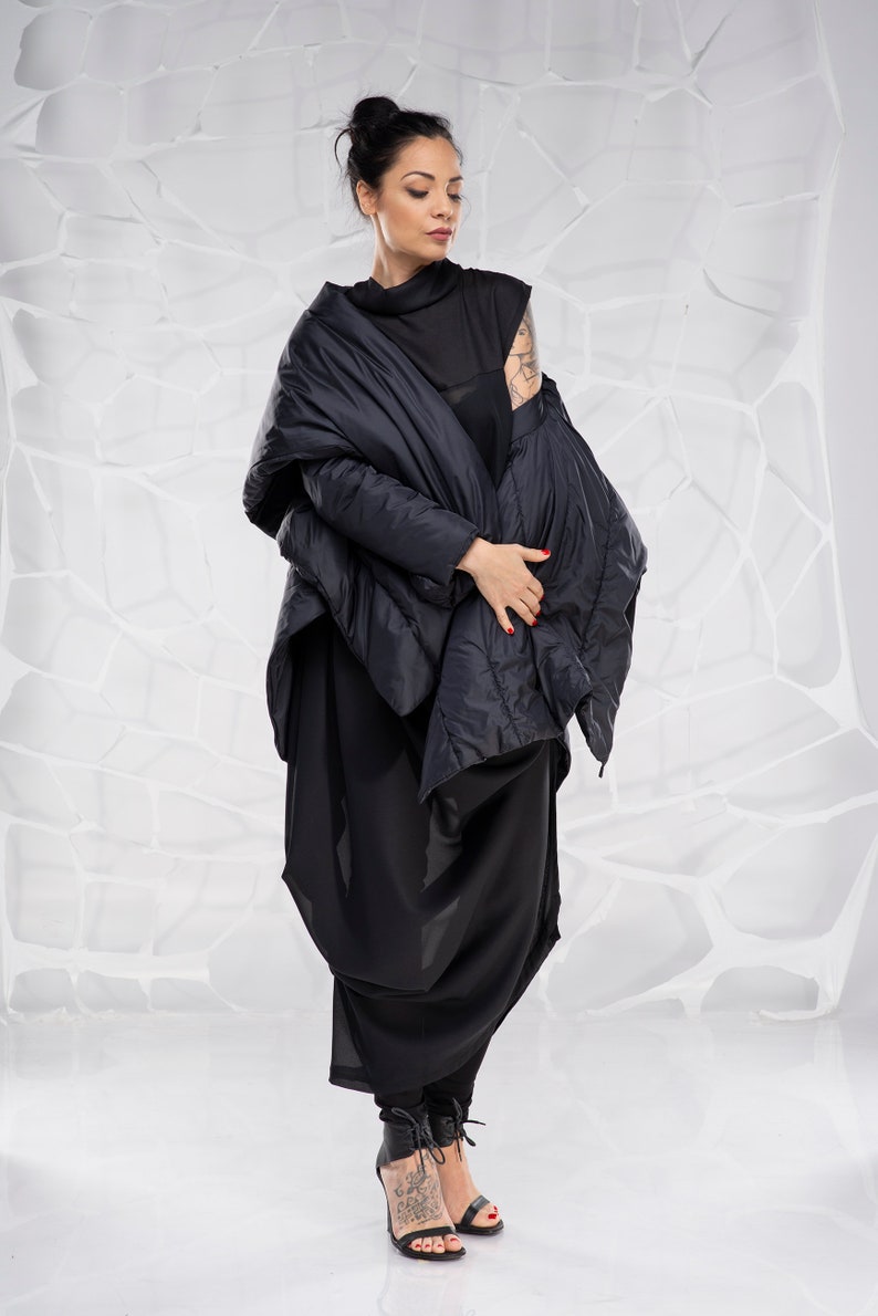 May include: A woman wearing a black, quilted jacket with a long, flowing skirt. She is standing in front of a white background.