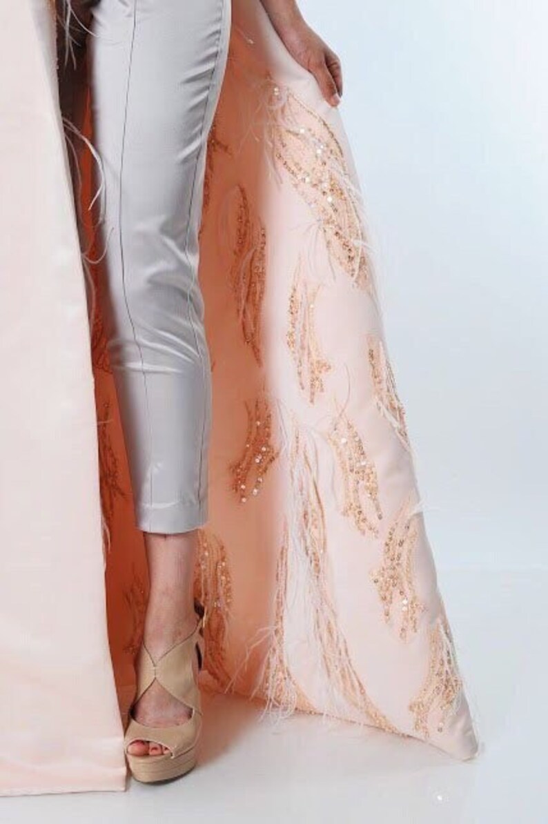 May include: A long, flowing pink cape with gold and white embroidery and feather trim. The cape is worn over a pair of light gray pants.