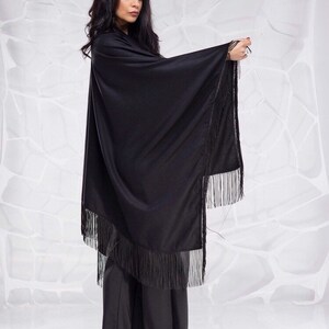 May include: A black shawl with fringe trim. The shawl is draped over a person's shoulders and arms. The person is wearing black pants and black heels.