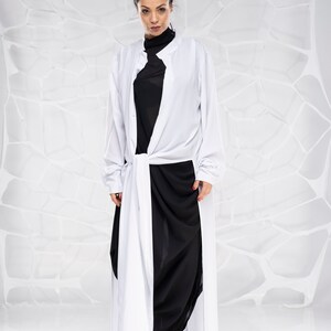 May include: A woman wearing a white long-sleeved shirt with a black shirt underneath and black pants. The shirt is tied at the waist and the pants are loose fitting. She is wearing black heels.