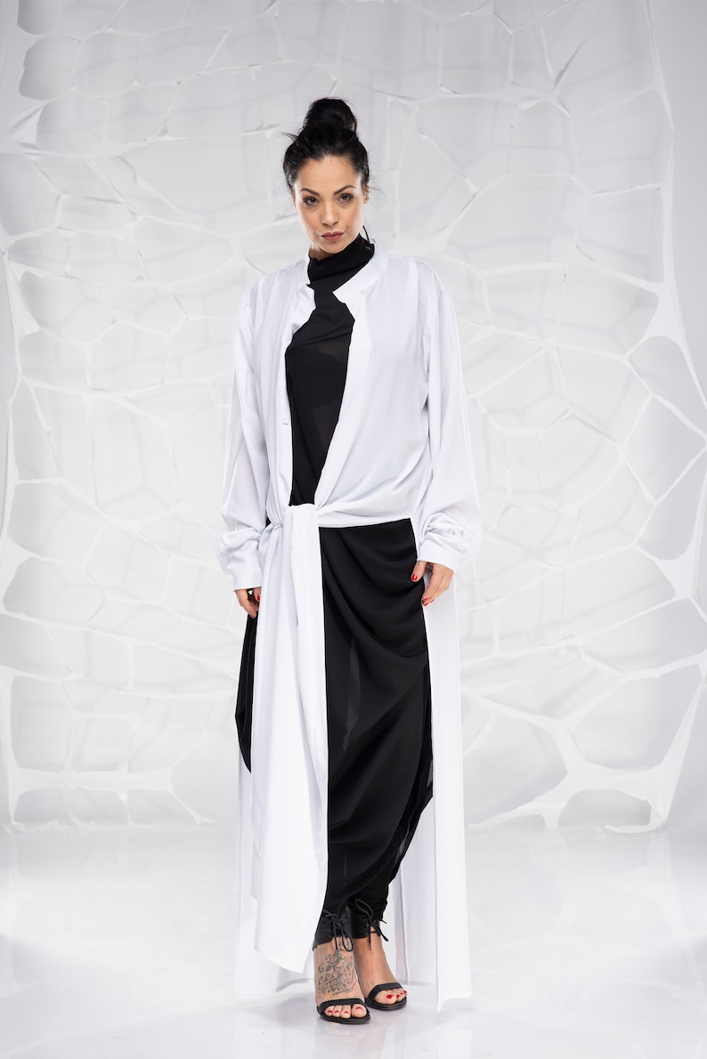 May include: A woman wearing a white long-sleeved shirt with a black shirt underneath and black pants. The shirt is tied at the waist and the pants are loose fitting. She is wearing black heels.