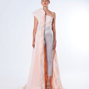 May include: A woman wearing a one-shoulder, blush pink gown with a long, feathered train. The gown has a fitted bodice and a flowing skirt. The woman is wearing silver pants underneath the gown.