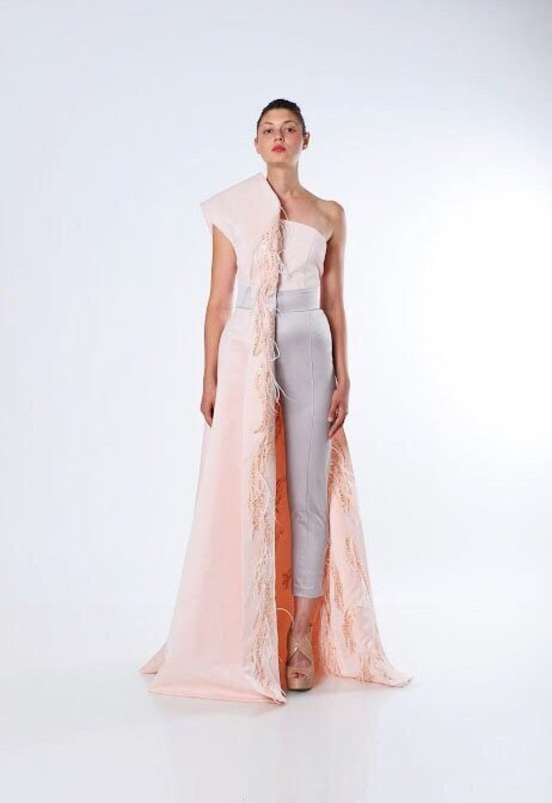 May include: A woman wearing a one-shoulder, blush pink gown with a long, feathered train. The gown has a fitted bodice and a flowing skirt. The woman is wearing silver pants underneath the gown.