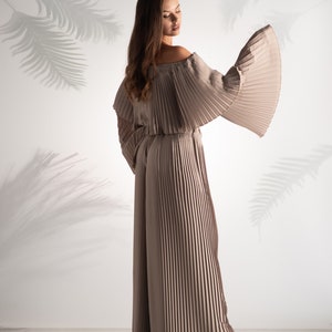 May include: A woman wearing a long, pleated, off-the-shoulder jumpsuit in a light brown color. The jumpsuit has wide, flowing sleeves and is made of a soft, flowing fabric.