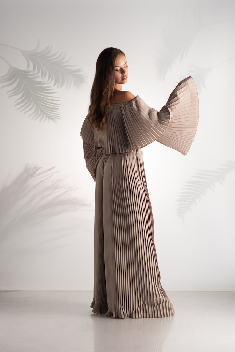 May include: A woman wearing a long, pleated, off-the-shoulder jumpsuit in a light brown color. The jumpsuit has wide, flowing sleeves and is made of a soft, flowing fabric.