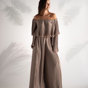 May include: A woman wearing a light brown off-the-shoulder top and matching wide-leg pants. The top and pants are made of pleated fabric.