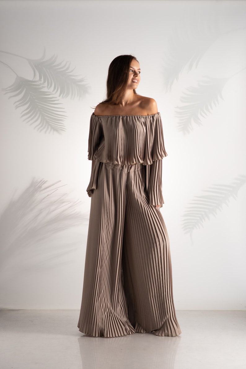 May include: A woman wearing a light brown off-the-shoulder top and matching wide-leg pants. The top and pants are made of pleated fabric.