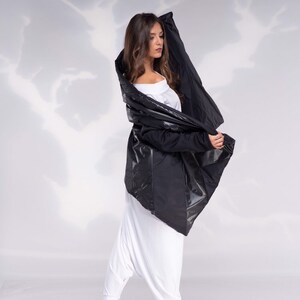 K&ouml;nnte beinhalten: Eine Frau tr&auml;gt eine schwarze, asymmetrische Jacke mit einem wei&szlig;en Oberteil und einer wei&szlig;en Hose. Sie tr&auml;gt schwarze Stiefel und steht auf einem wei&szlig;en Hintergrund.