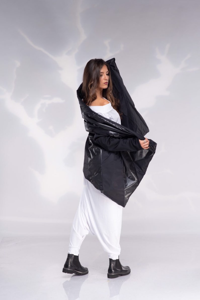 K&ouml;nnte beinhalten: Eine Frau tr&auml;gt eine schwarze, asymmetrische Jacke mit einem wei&szlig;en Oberteil und einer wei&szlig;en Hose. Sie tr&auml;gt schwarze Stiefel und steht auf einem wei&szlig;en Hintergrund.