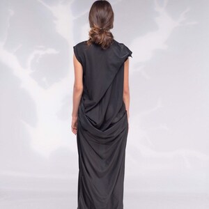 May include: A black, draped maxi dress with a unique asymmetrical design. The dress features a flowing silhouette and is paired with red high heels. The model's hair is styled in a bun. The background is a soft, white gradient.