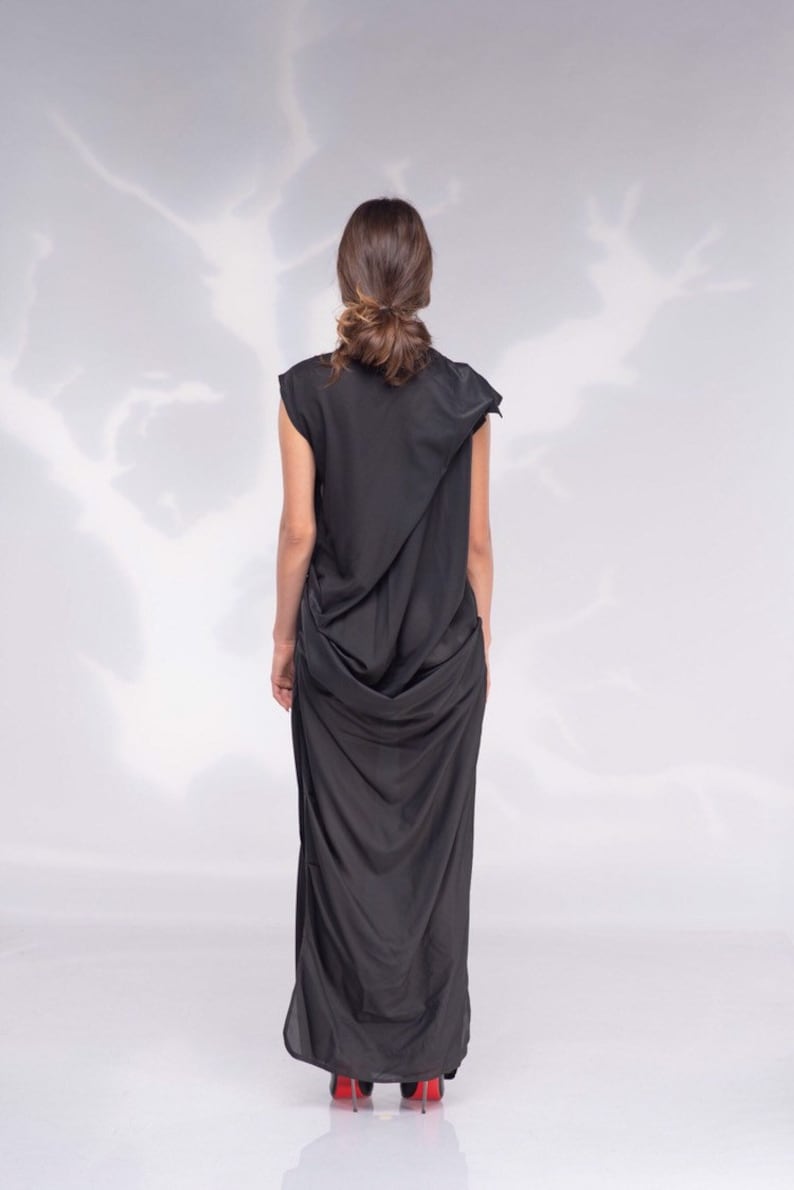 May include: A black, draped maxi dress with a unique asymmetrical design. The dress features a flowing silhouette and is paired with red high heels. The model's hair is styled in a bun. The background is a soft, white gradient.