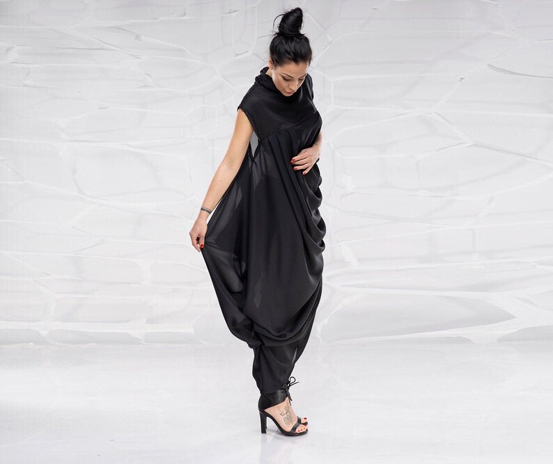 May include: A black, flowing, sleeveless dress with a high neck and a gathered, draped design. The dress is worn by a person with dark hair and a bun.