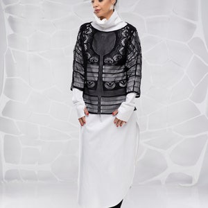 May include: A woman wearing a black lace top over a white long-sleeved shirt and black pants. She is standing in front of a white textured background.