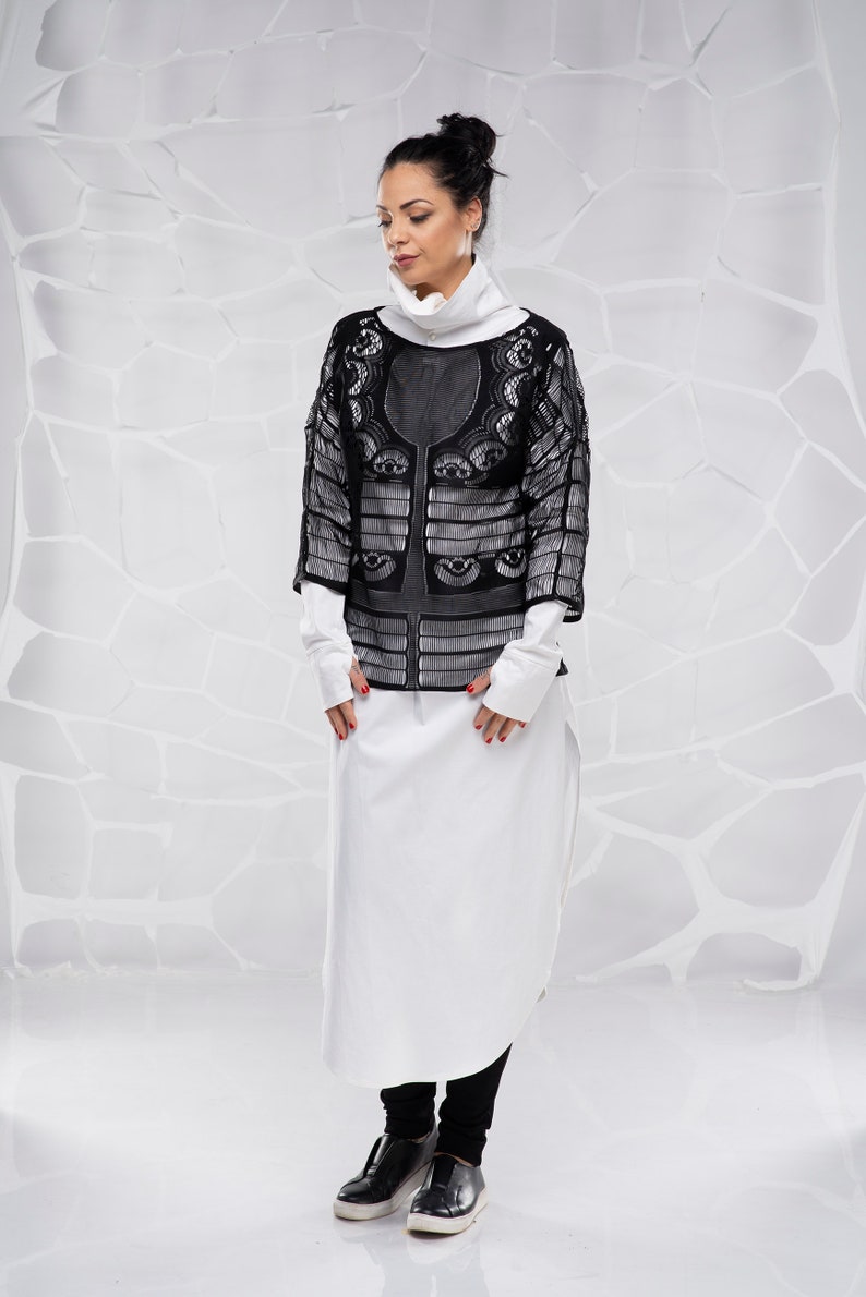May include: A woman wearing a black lace top over a white long-sleeved shirt and black pants. She is standing in front of a white textured background.