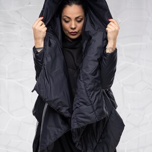 May include: A black hooded puffer jacket with a long, asymmetrical hemline. The jacket is made of a quilted fabric and has a relaxed fit.