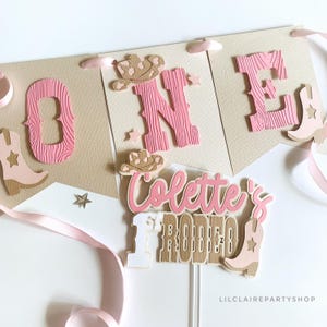 May include: A banner and cake topper set with a western theme. The banner spells "ONE" with pink wooden letters and cowboy boot accents. The cake topper reads "Colette's Rodeo" with a cowboy hat and boot. Pink and tan colors.