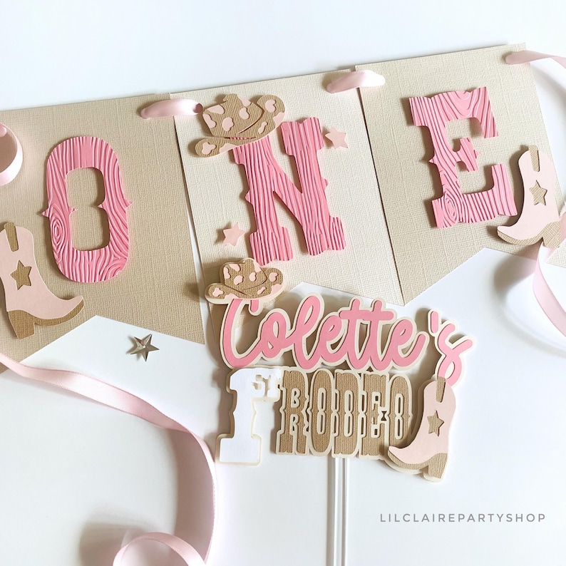 May include: A banner and cake topper set with a western theme. The banner spells "ONE" with pink wooden letters and cowboy boot accents. The cake topper reads "Colette's Rodeo" with a cowboy hat and boot. Pink and tan colors.