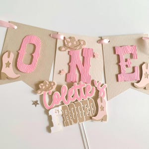 May include: A banner and cake topper set with a western theme. The banner spells "ONE" with pink letters and cowboy boot accents. The cake topper reads "Colette's Rodeo" in pink and tan. The set includes pink ribbons and star accents.