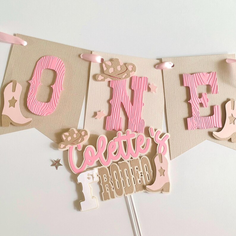 May include: A banner and cake topper set with a western theme. The banner spells "ONE" with pink letters and cowboy boot accents. The cake topper reads "Colette's Rodeo" in pink and tan. The set includes pink ribbons and star accents.