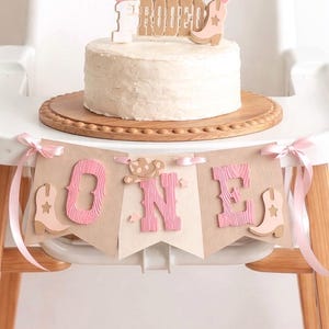 May include: A white frosted cake on a wooden board with a cake topper reading "Cowgirl Rodeo" and a banner below spelling "ONE" in pink letters, decorated with light pink cowboy boots and a hat, tied with pink ribbons on the sides.