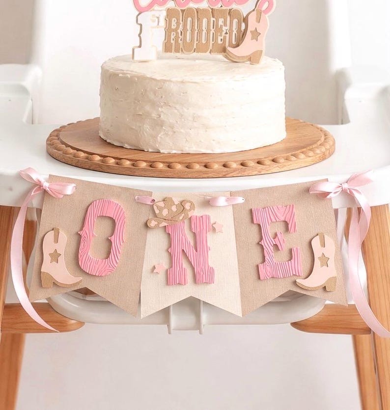 May include: A white frosted cake on a wooden board with a cake topper reading "Cowgirl Rodeo" and a banner below spelling "ONE" in pink letters, decorated with light pink cowboy boots and a hat, tied with pink ribbons on the sides.