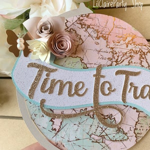 May include: A light pink and brown globe with a white banner that says "Time to Travel" in gold glitter. The globe is decorated with paper flowers and a butterfly.