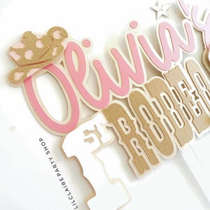 May include: A layered cake topper with the name "Olivia" in pink script, a tan cowboy hat, and the words "1st Birthday" in a tan and white font. The text "LILCLAIRE PARTY SHOP" is printed on the side.