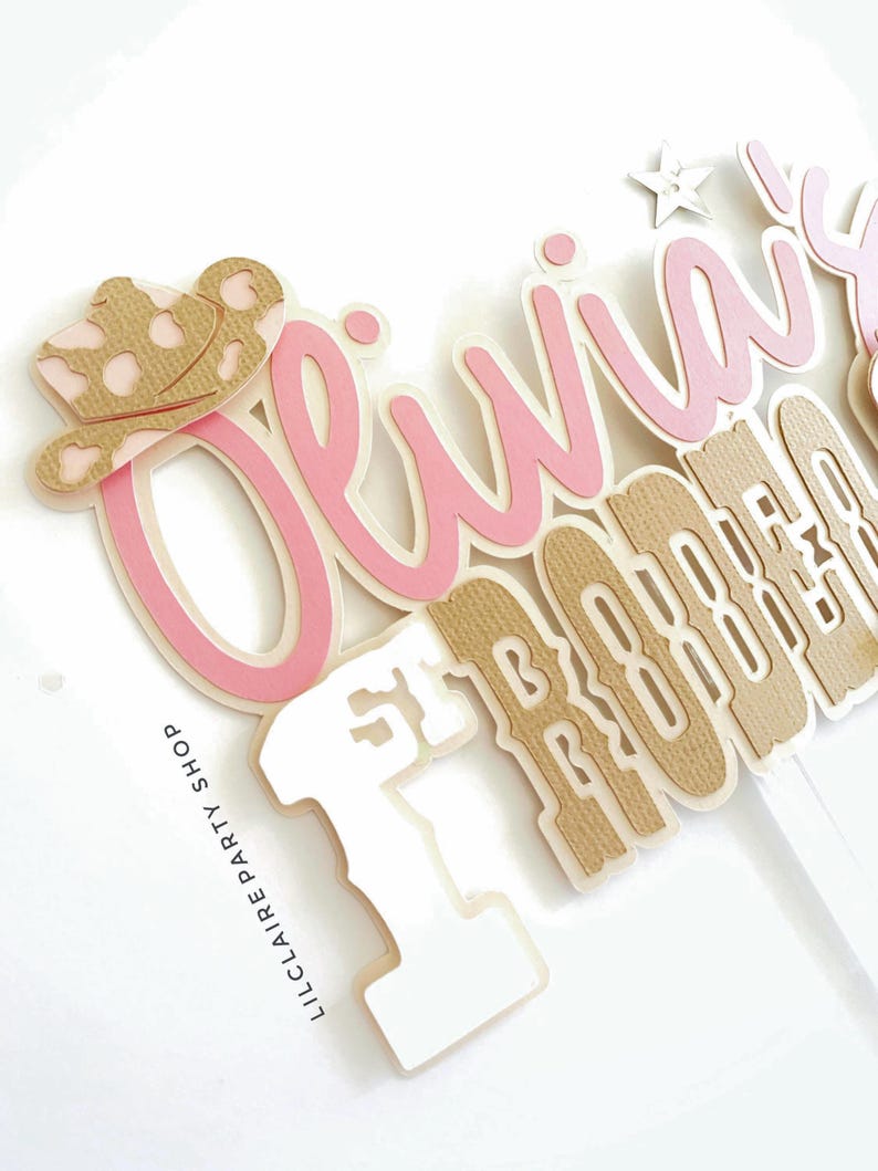 May include: A layered cake topper with the name "Olivia" in pink script, a tan cowboy hat, and the words "1st Birthday" in a tan and white font. The text "LILCLAIRE PARTY SHOP" is printed on the side.