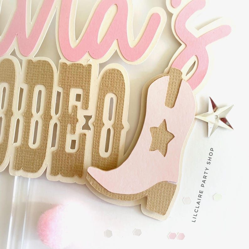 May include: A layered paper cake topper featuring the text "MA'S RODEO" in pink and tan textured cardstock. The design includes a light pink cowboy boot with a star cutout, a small silver star accent, and the text "LILCLAIRE PARTY SHOP".