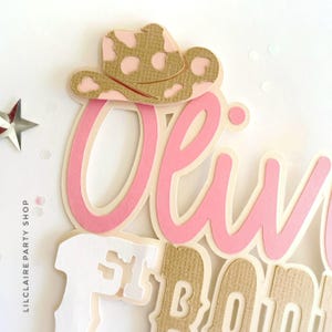 May include: A layered paper cake topper with the name "Olivia" in pink script, accented with a tan and pink cowboy hat. The words "First Birthday" are also visible. The design is set against a white background with a star and the text "LILCLAIRE PARTY SHOP".