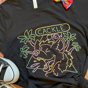 May include: Black t-shirt with a neon green, pink, and yellow graphic of a cartoon wolf howling with palm trees and the text "CACKLE CREW".