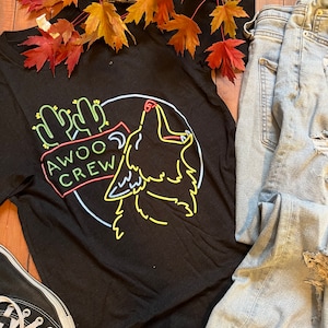 May include: Black t-shirt with a neon green, yellow, and red graphic of a howling wolf with cacti and the text "AWOO CREW".
